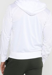 Vit hoodie med en texturerad yta, utrustad med en dragsko i huvan och ribbad nederkant. Baksidan visar ett subtilt mönster över tyget.