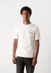 Krämfärgad bomullströja med en färgglad grafik på bröstet och texten "Skate & Surf," matchad med mörk denimjeans.