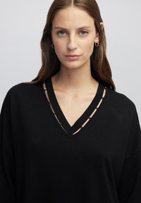 Schwarzer Pullover mit V-Ausschnitt-Design, ausgestattet mit Netz-Ausschnitten und metallischen Akzenten. Der Stoff hat eine glatte Textur und einen lockeren Schnitt.