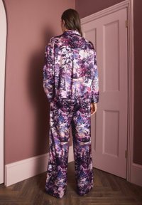 Sieviete stāv pretim mauve krāsas sienai un durvīm, ģērbusies vaļīgā, saskaņotā jaku un biksēs ar purpura, rozā un baltu abstraktu ziedu rakstu.