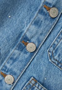 Denimtyg i ljusblått med koppartråd, prytt med silverfärgade metallknappar och små dekorativa detaljer.