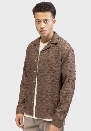 Brown textured shirt featuring a wavy pattern, buttoned front, and collar. Worn over a light-colored knit top, with visible stitching details.