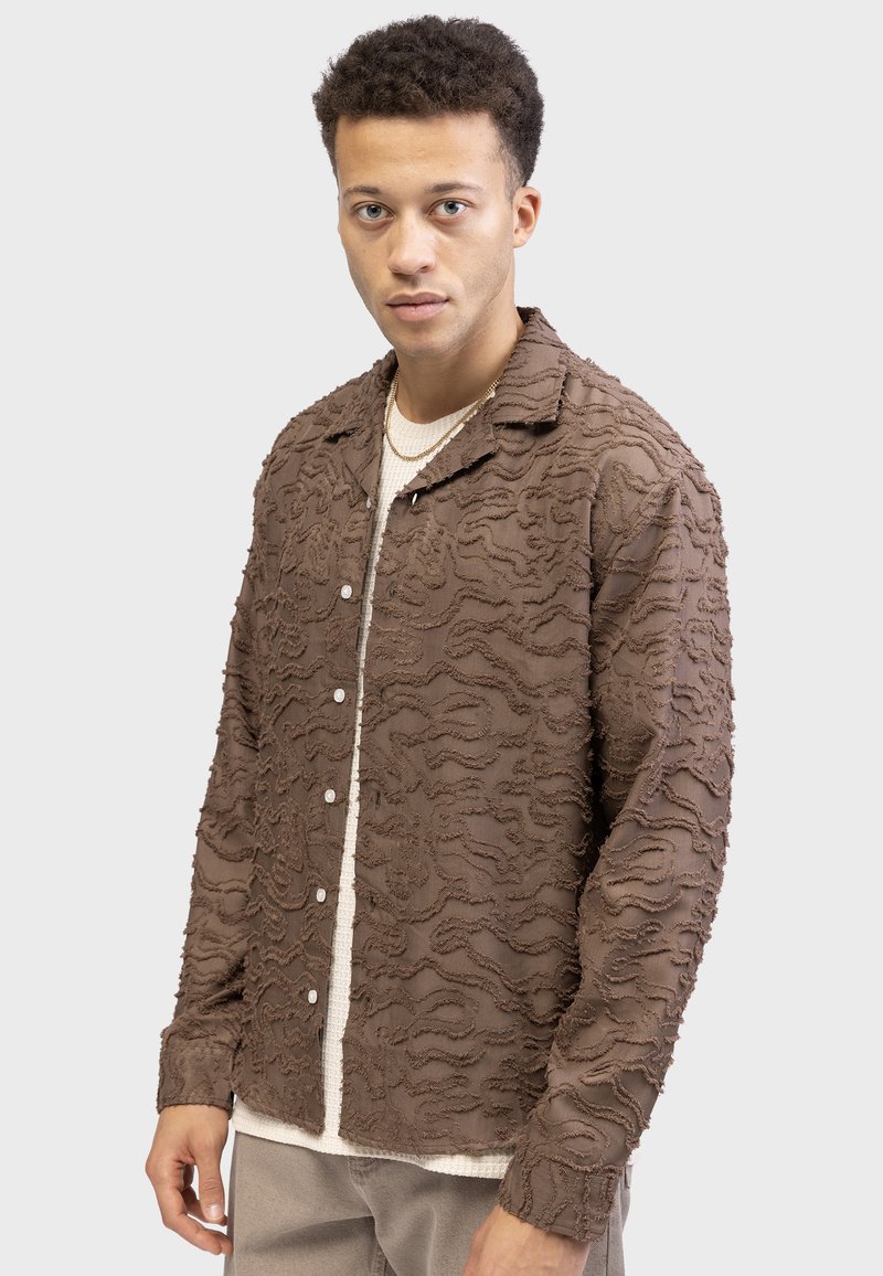 Brown textured shirt featuring a wavy pattern, buttoned front, and collar. Worn over a light-colored knit top, with visible stitching details.