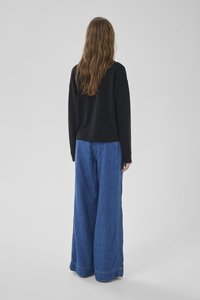 Persona con capelli castani lunghi che indossa una maglia nera a maniche lunghe e jeans blu a gamba larga, vista di spalle su uno sfondo grigio uniforme.