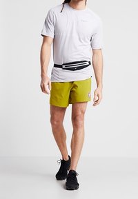 Hellgraues sportliches T-Shirt, gelbe Shorts mit Logo, schwarze Bauchtasche und schwarze Sneakers. Freizeit-Sportbekleidung mit feuchtigkeitsableitendem Design.