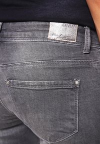 Jeans de denim negro con un lavado medio, que presentan una etiqueta de marca de cuero y herrajes de plata en los bolsillos traseros. Diseño clásico de cinco bolsillos.