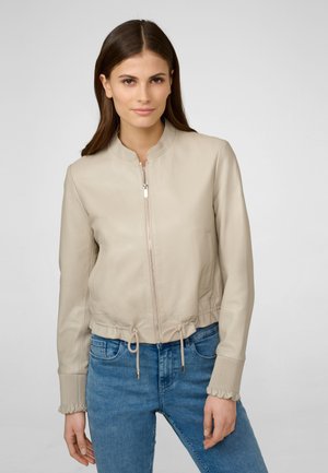 Beige leren bomberjack met een ritssluiting aan de voorkant, ronde kraag en een trekkoord aan de onderkant. Voorzien van ribbels bij de manchetten met gerafelde randen. Jeans zichtbaar eronder.