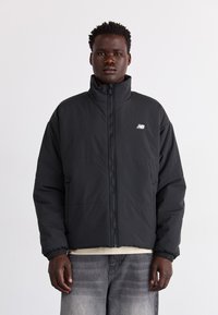 Svart isolerad jacka med hög krage, dragkedjestängning och elastiska ärmslut. Har en liten grå New Balance-logotyp på bröstet.
