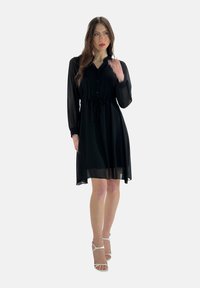 Abito camicia nero realizzato in materiale trasparente con maniche lunghe, chiusura con bottoni frontale, vita cintrata e gonna fluida fino a metà coscia.