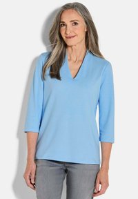 Lichtblauwe V-hals blouse van zachte stof, met driekwartmouwen en een relaxte pasvorm, gecombineerd met grijze broek.