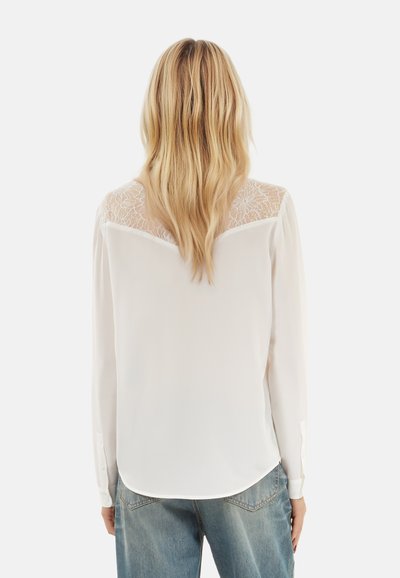 Blusa bianca con retro in pizzo trasparente, maniche lunghe, polsini con bottoni e orlo arrotondato. Indossata con jeans blu dalla vestibilità comoda.