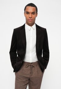 Brun kordflorblazer med notch-lapel, parat med en vit skjorta med knappar. Bruna skräddarsydda byxor har en dragsko vid midjan och en texturerad tyg.