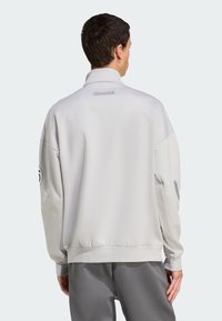 Lys grå sportssweatshirt med høj krave, elastiske manchetter og et tekstureret stof. Har branding på ryggen og ærme detaljer.