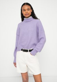 Sieviete ar cirtainiem matiem, valkājot lavandas krāsas augstu apkaklīti džemperi, baltas šortus ar melnu jostu, stāvot pret vienkāršu gaišu fonu.