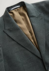 Blazer vert foncé en tissu texturé ; il présente un revers cranté et une doublure en satin marron à l'intérieur. Comprend des détails de couture subtils.