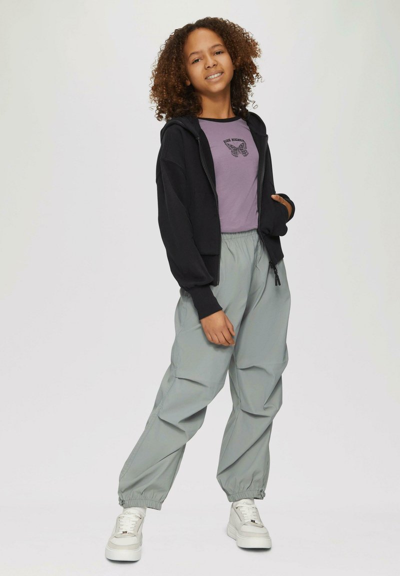 Schwarze Zip-Up-Hoodie über einem lila T-Shirt mit Schmetterlingsdruck, kombiniert mit hellgrauen, lockeren Hosen. Weiße Sneakers runden den Look ab.