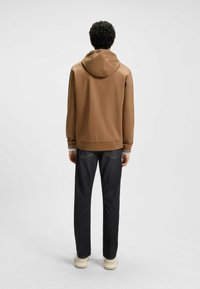 Brown hooded sweatshirt with a smooth texture, elastic cuffs with white and black stripes, paired with dark denim pants and light-colored shoes.