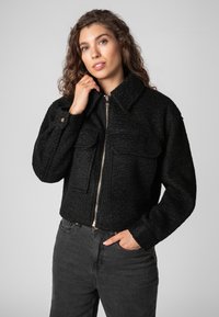 Black cropped jacket, textured fabric, large chest pockets, front zipper, and a shirt collar. Worn with black jeans.