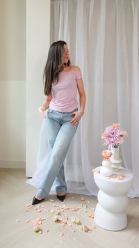 Rosa off-shoulder topp, ljusblå jeans och svarta spetsiga klackar. Vit skulptural bord med blommor och blad på golvet. Neutral bakgrund.