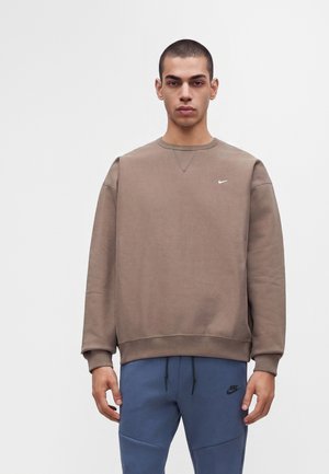Joven con el pelo corto vistiendo una sudadera marrón de Nike y pantalones deportivos azules de Nike, de pie frente a un fondo blanco liso.