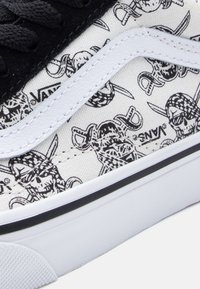 Μαύρο καμβά αθλητικό παπούτσι με λευκά ράμματα, εκτυπωμένο σχέδιο πειρατικού κρανίου σε κρεμ φόντο και λευκή καουτσούκ σόλα με μαύρη ρίγα.