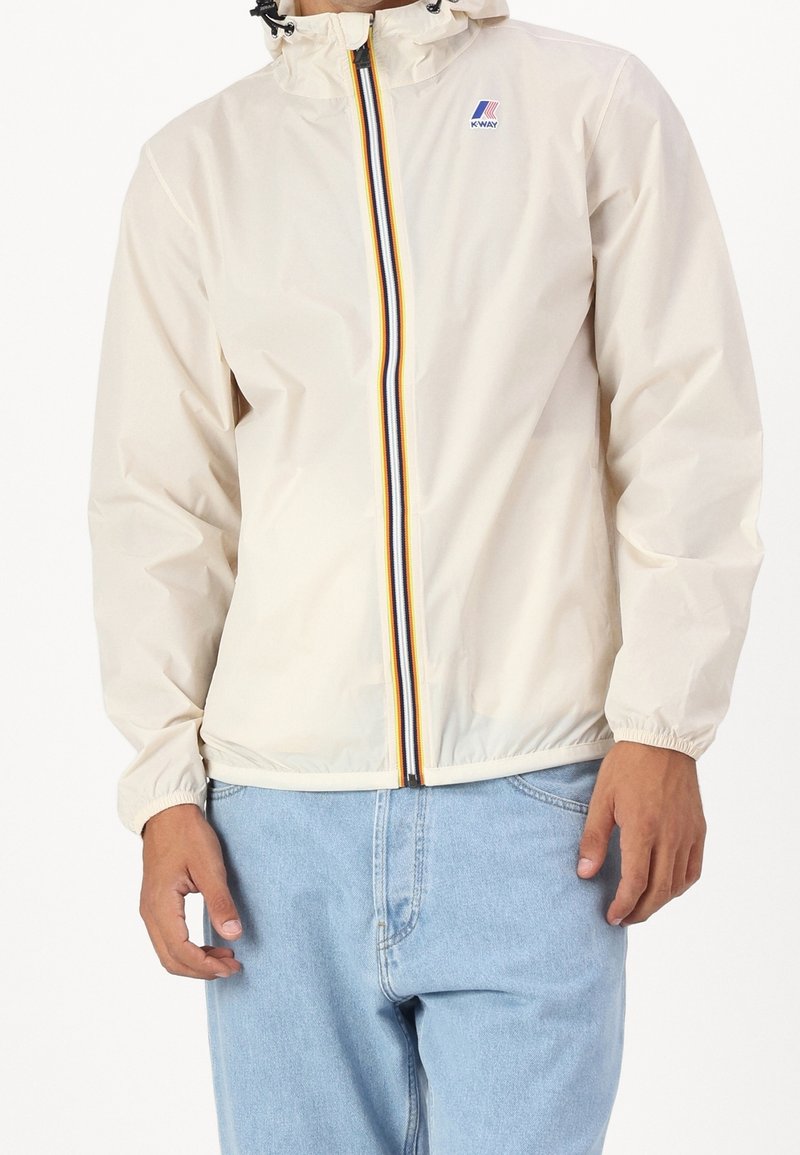 Světle béžová nepromokavá bunda s kapucí, vybavená předním zavíráním na zip zdobeným černými, žlutými a červenými pruhy. Elastické manžety a spodní lem.