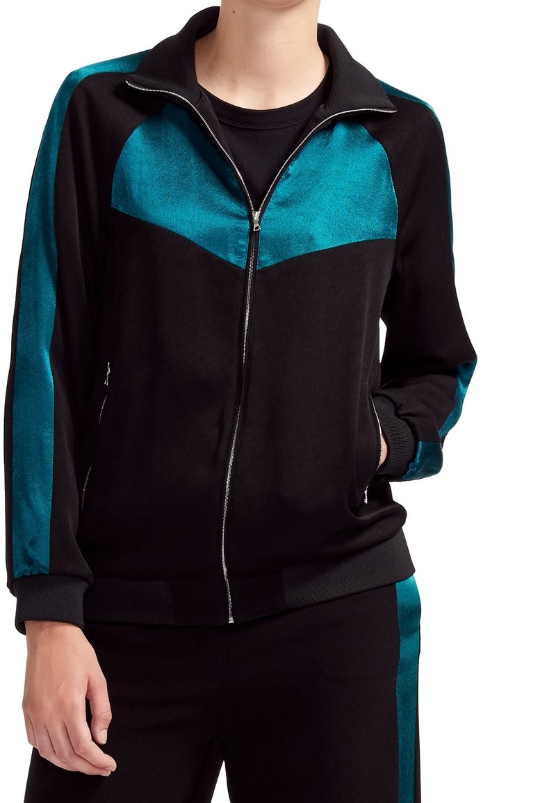 Personne portant une veste noire zippée avec des panneaux bleu sarcelle brillants sur la poitrine et les manches, ainsi qu'un pantalon noir assorti avec des bandes bleu sarcelle sur les côtés.