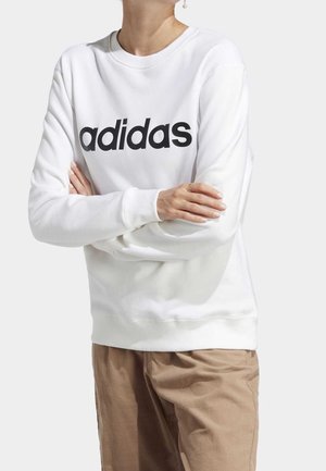 Osoba w białej bluzie z czarnym logo "adidas" na piersi i beżowych spodniach, z założonymi skrzyżowanymi rękoma.
