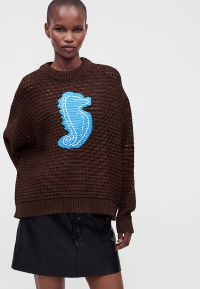 Personne portant un pull en maille marron ample avec un grand motif hippocampe bleu et une jupe en cuir noire boutonnée sur un fond uni.