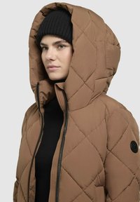 Hnedá prešívaná puffer bunda s veľkou kapucňou, čiernym zipsom a kruhovou nášivkou na ramene. Textúrovaná tkanina, nosená cez čierny top.
