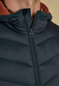 Zbliżenie mężczyzny w czarnej pikowanej kurtce na zamek błyskawiczny, noszącego czerwoną koszulę w kratę, z widocznymi detalami kołnierza i zamka kurtki.