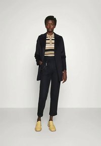 Jūras spēku mētelis, svītrainā polo krekls ar beidžu un jūras spēku toņu, tumši bikses un dzeltenas čības. Gluda tekstūra, mūsdienu griezums, minimāli aparatūras akcenti.