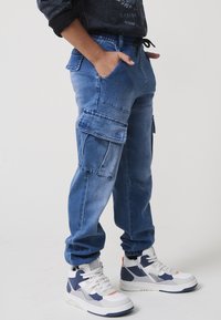 Világoskék cargo nadrág, több zsebbel, húzózsinóros derékkal, szűkített lábakkal és megfakult farmer textúrával, fehér és tengerészkék sportcipővel párosítva.