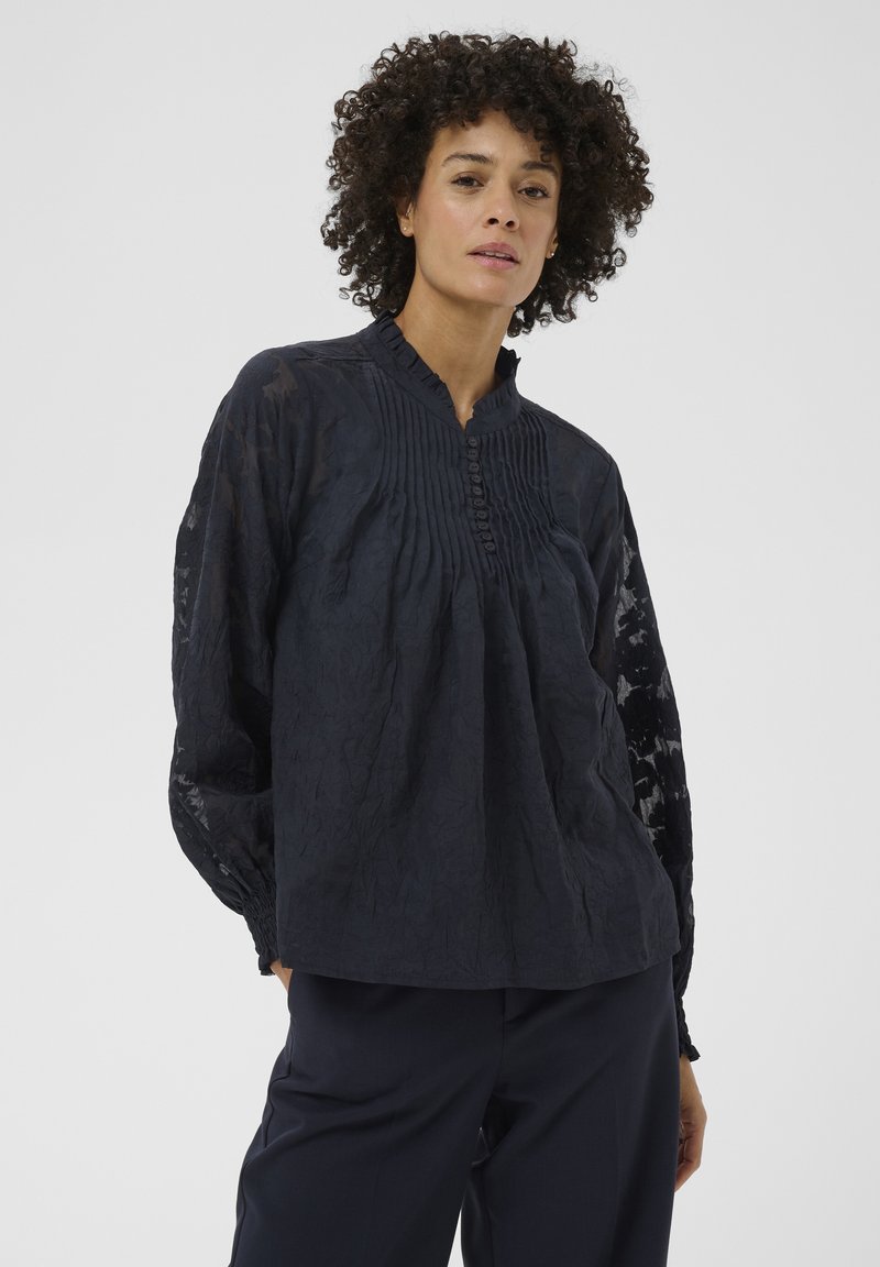 Femme aux cheveux bouclés portant un chemisier à manches longues bleu marine foncé avec des plis et des boutons sur le devant, assorti à un pantalon foncé.