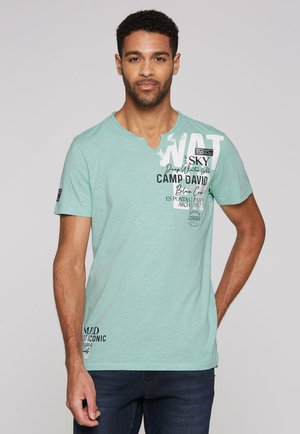 T-shirt in cotone verde menta con scollo a V, caratterizzato da testi e disegni stampati in bianco e nero, maniche corte e una vestibilità ampia.