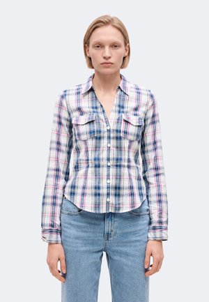 Femme aux cheveux blonds courts portant une chemise à carreaux bleue, blanche et rose boutonnée et un jean bleu clair, regardant devant elle avec une expression neutre.