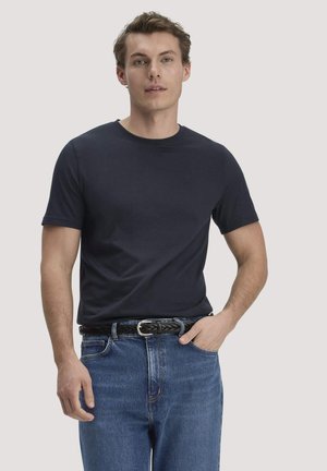 Junger Mann trägt ein schlichtes marineblaues T-Shirt, das in blaue Jeans mit geflochtenem braunem Gürtel gesteckt ist, und steht vor einem hellgrauen Hintergrund.