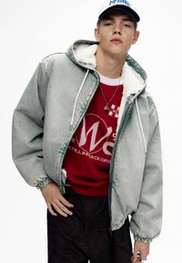 Jachetă din denim de culoare albastru deschis, cu căptușeală din fleece alb, manșete elastice și glugă cu șnur, purtată peste un hanorac grafic roșu și pantaloni cu model.