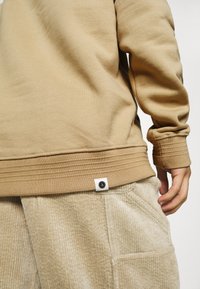 Felpa in cotone beige con polsini e orlo a costine, dotata di etichetta. Abbinata a pantaloni in corduroy beige chiaro, che mostrano una texture verticale.