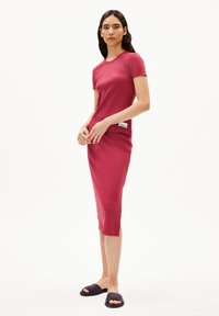 Enganliegendes, geripptes Kleid in Dunkelrot, mit kurzen Ärmeln, knielang und seitlichen Schlitzen. Getragen mit dunklen Slipper-Sandalen. Minimalistisches Etikettendetail.