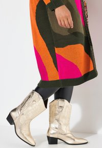 Bottines de cowboy dorées avec des motifs de feuilles en relief et un bout pointu, associées à une robe en tricot colorée avec des motifs audacieux en orange et rose.