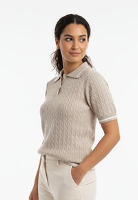 Beżowa polo z krótkim rękawem wykonana z dzianiny w warkocze, z jasnoszarym kołnierzem. Posiada ściągacze na mankietach oraz luźny krój. Gładka tekstura.