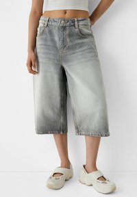 Persona vistiendo pantalones cortos de mezclilla gris desgastados hasta las rodillas y zapatillas blancas gruesas sin cordones, de pie contra un fondo blanco.