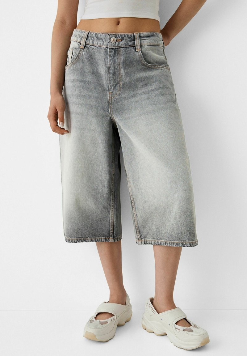 Persona vistiendo pantalones cortos de mezclilla gris desgastados hasta las rodillas y zapatillas blancas gruesas sin cordones, de pie contra un fondo blanco.