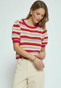 Kurzarmpullover aus Strick mit mehrfarbigen Streifen (rot, lila, gelb). Hat einen gewellten Ausschnitt und strukturierten Stoff. Getragen mit beigen Hosen.