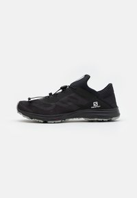 Chaussure de sport noire avec tige en mesh, un système de laçage rapide et une semelle texturée ; présente un logo sur le côté et à l'arrière.