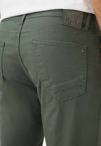 Donkergroene broek van een gestructureerde stof. Bevat een leren logo patch en twee achterzakken met gebogen flappen. Stiksel is zichtbaar.