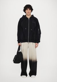 Hanorac negru cu fermoar, realizat dintr-un material texturat, având buzunare mari și un logo. Pantaloni cu gradient bej-negru, cu detalii uzate, asortați cu o geantă neagră.