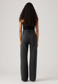 Jeans noirs taille haute et à jambes larges avec une texture en denim et un léger délavage. Comprend des poches arrière et une petite étiquette de marque rouge sur la taille.