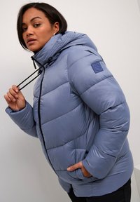 Giubbotto imbottito di colore azzurro chiaro, dotato di cappuccio, coulisse, zip anteriore e tasche laterali. Texture liscia con design trapuntato.